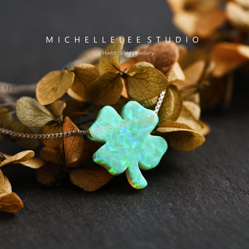 May include: A silver necklace with a four-leaf clover pendant. The pendant has an iridescent, light green finish with blue and orange hues. The necklace is from Michelle Lee Studio, which is described as handcrafted jewellery.