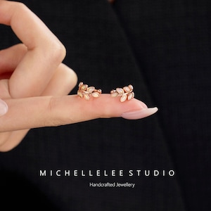 Puede incluir: Un par de pendientes ear cuff de oro rosa con forma de delicadas ramas de hojas, cada uno con peque&ntilde;as piedras ovaladas iridiscentes engastadas a lo largo del metal. El texto MICHELLELEE STUDIO Handcrafted Jewellery aparece abajo.