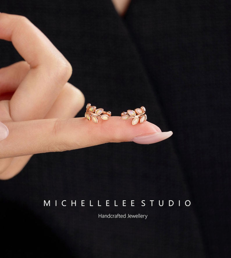 Puede incluir: Un par de pendientes ear cuff de oro rosa con forma de delicadas ramas de hojas, cada uno con peque&ntilde;as piedras ovaladas iridiscentes engastadas a lo largo del metal. El texto MICHELLELEE STUDIO Handcrafted Jewellery aparece abajo.