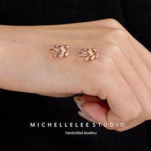 Puede incluir: Un par de pendientes de oro rosa con forma de hoja, cada uno con peque&ntilde;os &oacute;palos ovalados iridiscentes engastados a lo largo del marco de metal. El texto MICHELLELEE STUDIO Handcrafted Jewellery est&aacute; impreso en blanco en la parte inferior.