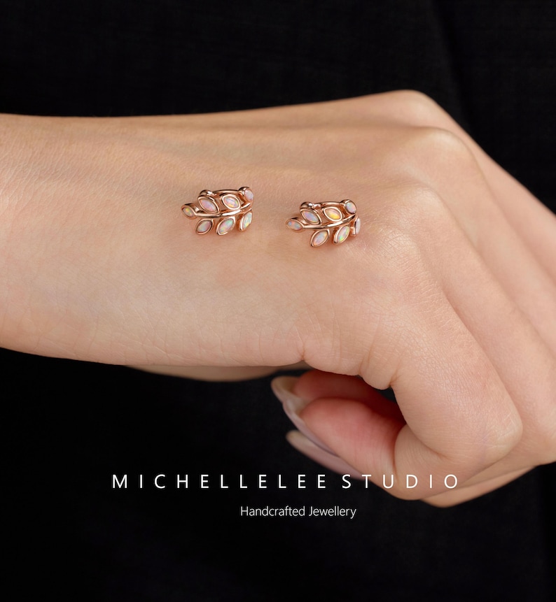 Puede incluir: Un par de pendientes de oro rosa con forma de hoja, cada uno con peque&ntilde;os &oacute;palos ovalados iridiscentes engastados a lo largo del marco de metal. El texto MICHELLELEE STUDIO Handcrafted Jewellery est&aacute; impreso en blanco en la parte inferior.