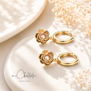 May include: Two gold-coloured hoop earrings. Each earring has a flower-shaped charm with a pearl centre. The earrings are presented on a white surface with soft lighting and floral details. The brand name "mi Chellelee" is visible.