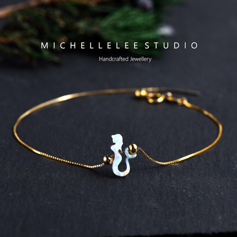 Puede incluir: Una delicada pulsera dorada con un dije de caballito de mar en tonos blancos y azules iridiscentes. El dije est&aacute; acentuado con dos peque&ntilde;as cuentas doradas. De MICHELLELEE STUDIO, Handcrafted Jewellery.