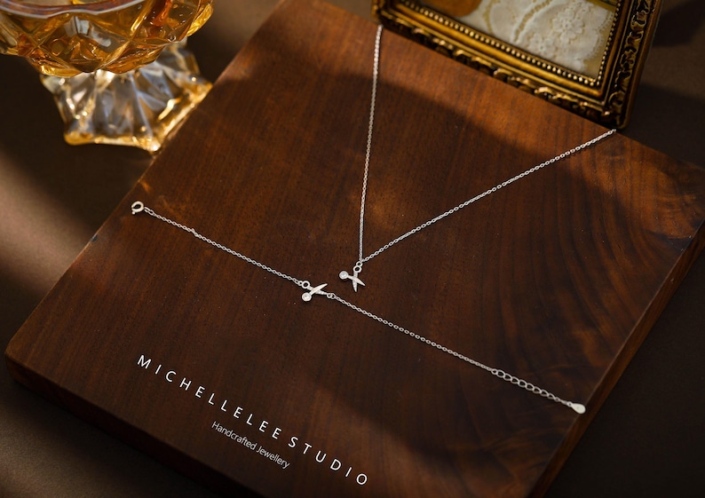May include: A silver necklace and bracelet set with a delicate scissor charm. The jewelry is displayed on a wooden surface with the text "MICHELLELEE STUDIO Handcrafted Jewellery".