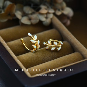 Puede incluir: Dos pendientes tipo ear cuff dorados con forma de ramas de hojas, cada uno con peque&ntilde;as piedras de &oacute;palo blanco ovaladas. El texto MICHELLELEE STUDIO Handcrafted Jewellery es visible en la parte inferior.