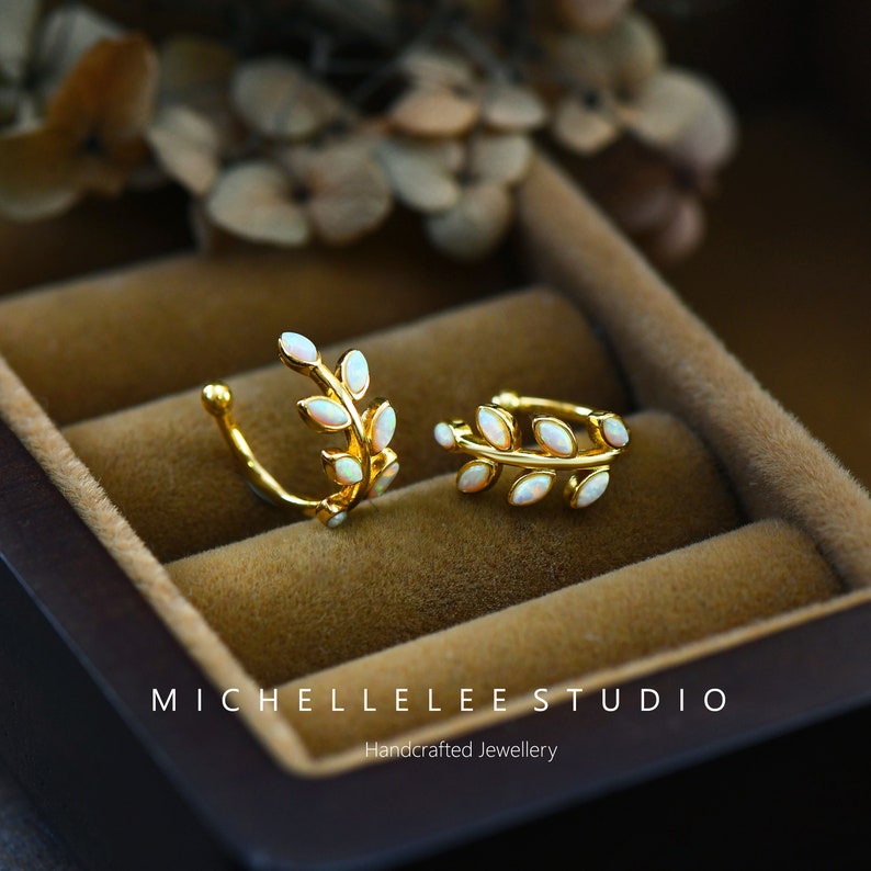 Puede incluir: Dos pendientes tipo ear cuff dorados con forma de ramas de hojas, cada uno con peque&ntilde;as piedras de &oacute;palo blanco ovaladas. El texto MICHELLELEE STUDIO Handcrafted Jewellery es visible en la parte inferior.