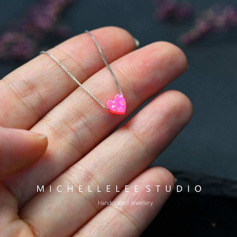 May include: A silver necklace with a small, pink heart-shaped pendant. The pendant has a textured finish. The necklace is shown on a hand. The text "MICHELLELEE STUDIO" and "Handcrafted Jewellery" are visible.