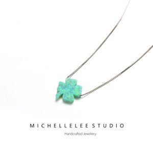 May include: A silver necklace with a four-leaf clover pendant in light blue and green. The pendant has an iridescent finish. The necklace is a delicate piece of jewellery. The text "MICHELLELEE STUDIO" and "Handcrafted Jewellery" are visible.