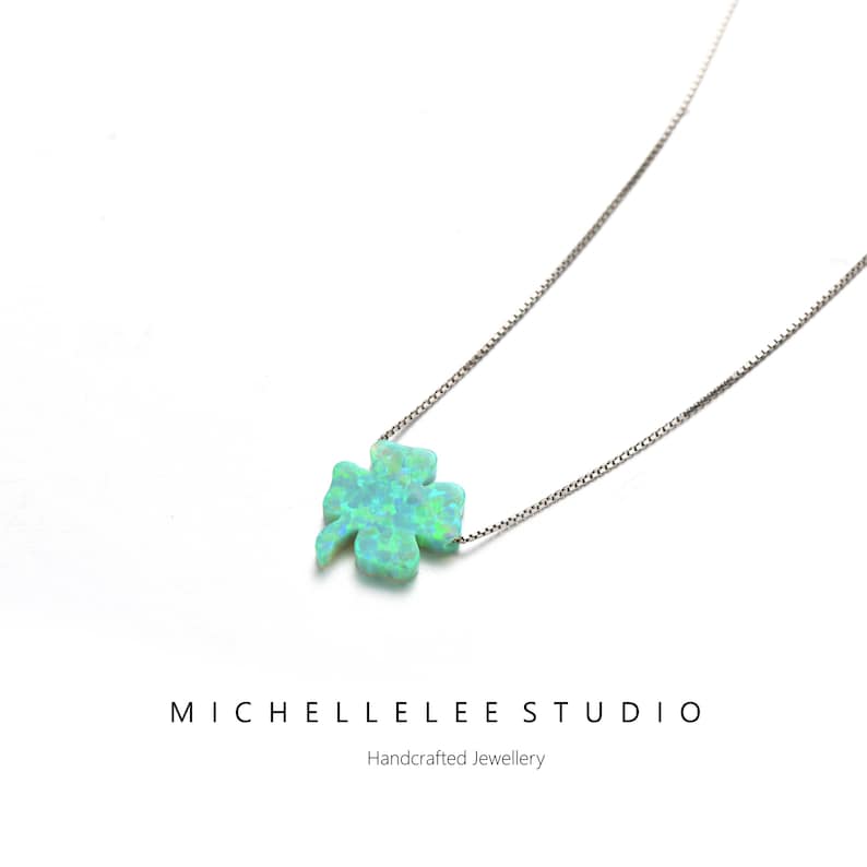 May include: A silver necklace with a four-leaf clover pendant in light blue and green. The pendant has an iridescent finish. The necklace is a delicate piece of jewellery. The text "MICHELLELEE STUDIO" and "Handcrafted Jewellery" are visible.