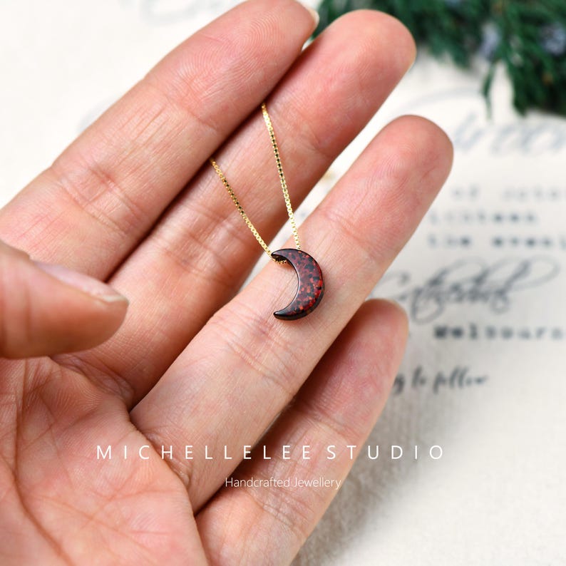 May include: A gold-colored chain necklace with a crescent moon pendant. The moon pendant is dark with red speckles. The necklace is displayed on a hand. The text "MICHELLELEE STUDIO Handcrafted Jewellery" is visible.