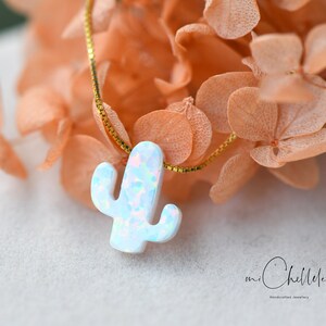 Op de afbeelding: Een delicate gouden ketting met een witte opaal cactus hanger. De hanger heeft een lichtblauwe en roze iriserende glans. De ketting wordt getoond op een bed van gedroogde bloemen.
