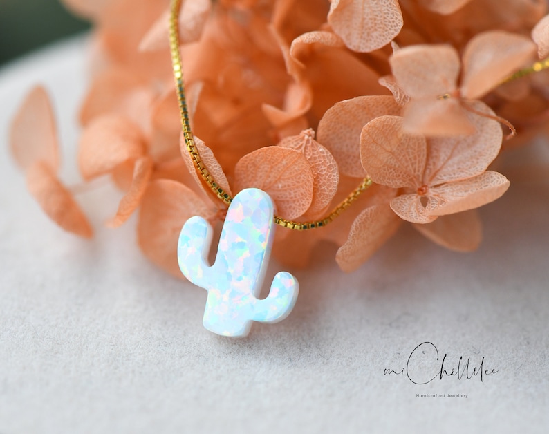 Op de afbeelding: Een delicate gouden ketting met een witte opaal cactus hanger. De hanger heeft een lichtblauwe en roze iriserende glans. De ketting wordt getoond op een bed van gedroogde bloemen.