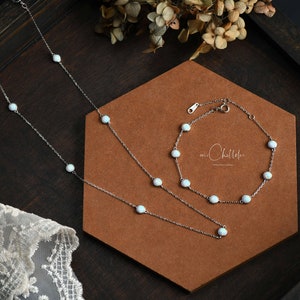 Pode incluir: Um conjunto de colar e pulseira de prata com pedras brancas semelhantes a opalas. As joias s&atilde;o exibidas em uma superf&iacute;cie marrom com um fundo de madeira escura. A pulseira e o colar t&ecirc;m um design delicado.