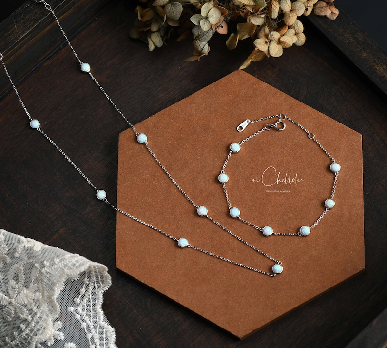 Pode incluir: Um conjunto de colar e pulseira de prata com pedras brancas semelhantes a opalas. As joias s&atilde;o exibidas em uma superf&iacute;cie marrom com um fundo de madeira escura. A pulseira e o colar t&ecirc;m um design delicado.