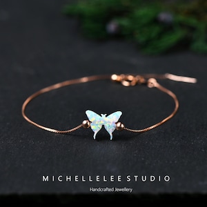 Pode incluir: Uma delicada pulseira em ouro rosa com um pingente de borboleta com asas iridescentes semelhantes a opala. A borboleta está centrada na corrente, ladeada por pequenas contas redondas. A pulseira é exibida em uma superfície escura. Texto: "MICHELLELEE STUDIO Handcrafted Jewellery."