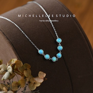 May include: A delicate silver necklace featuring seven round, light blue opal stones. The stones are evenly spaced along the chain. The necklace is displayed on a brown surface. Text on the image reads "MICHELLE LEE STUDIO Handcrafted Jewellery".
