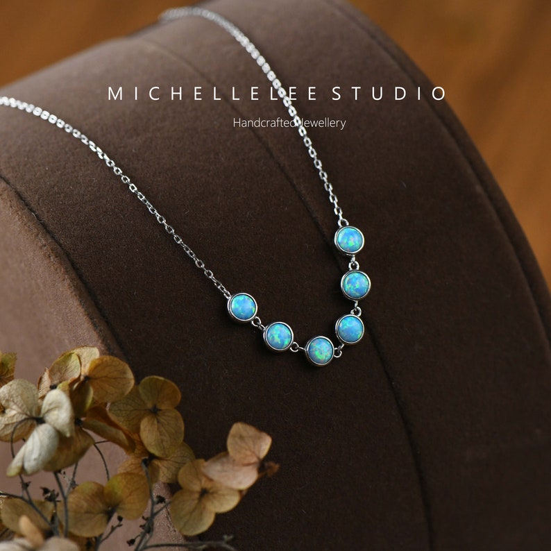 May include: A delicate silver necklace featuring seven round, light blue opal stones. The stones are evenly spaced along the chain. The necklace is displayed on a brown surface. Text on the image reads "MICHELLE LEE STUDIO Handcrafted Jewellery".