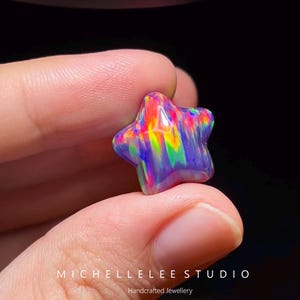 Pode incluir: Uma pedra preciosa de opala em forma de estrela com um jogo de cores vibrante, apresentando tons de vermelho neon, amarelo, verde e roxo profundo. O texto MICHELLELEE STUDIO Handcrafted Jewellery está impresso em branco na parte inferior.