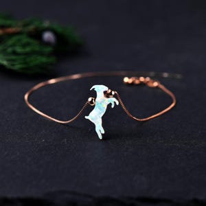 Op de afbeelding: Een delicate roségouden armband met een geitenvormige hanger met een glanzende, opalescente afwerking. De geit staat op zijn achterpoten. De armband heeft een fijne ketting en kleine, ronde kralen.