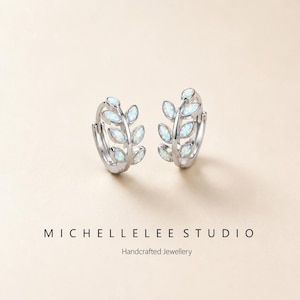 May include: A pair of silver hoop earrings with a leaf design. Each earring features a series of opal-like stones set in a leaf pattern. The earrings are displayed on a light beige background. The text "MICHELLELEE STUDIO" and "Handcrafted Jewellery" are below the earrings.