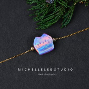 May include: A gold chain necklace with a cat-shaped pendant. The pendant is iridescent with pink, blue, and purple hues. The necklace has small gold beads on either side of the pendant. The text "MICHELLELEE STUDIO" and "Handcrafted Jewellery" are at the bottom.