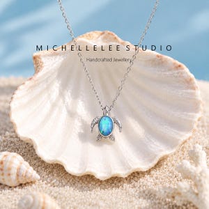 May include: A silver-toned necklace featuring a turtle pendant with an oval-shaped iridescent blue opal inlay. The text MICHELLELEE STUDIO Handcrafted Jewellery is visible above the pendant. The chain is a delicate silver-toned link style.