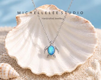 Może przedstawiać: Srebrzysty naszyjnik z zawieszką w kształcie żółwia z owalną, opalizującą niebieską wstawką z opalu. Tekst MICHELLELEE STUDIO Handcrafted Jewellery jest widoczny nad zawieszką. Łańcuszek wykonany jest z delikatnych, drobnych ogniw.