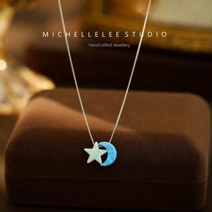 May include: A silver necklace with a blue opal crescent moon and a white star charm. The necklace is on a brown velvet box. The text "MICHELLELEE STUDIO Handcrafted Jewellery" is on the box.