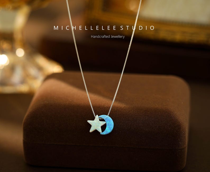 May include: A silver necklace with a blue opal crescent moon and a white star charm. The necklace is on a brown velvet box. The text "MICHELLELEE STUDIO Handcrafted Jewellery" is on the box.