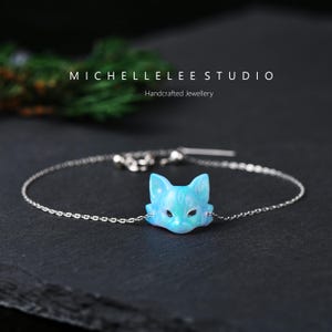 May include: A silver bracelet with a light blue fox head pendant. The fox head has a shimmering finish and dark eye details. The bracelet is from MICHELLELEE STUDIO, handcrafted jewellery.