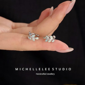 Puede incluir: Dos pendientes de presi&oacute;n plateados con forma de hoja, cada uno con peque&ntilde;as piedras ovaladas iridiscentes. El texto MICHELLELEE STUDIO Handcrafted Jewellery aparece en la parte inferior de la imagen.