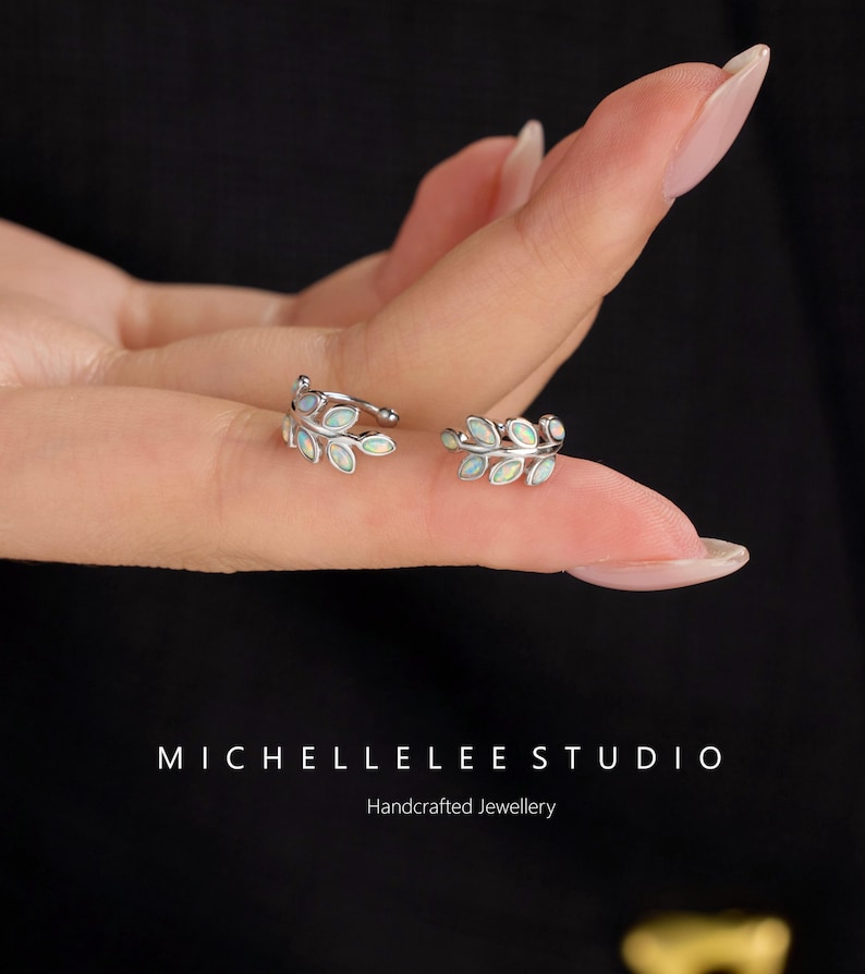 Puede incluir: Dos pendientes de presi&oacute;n plateados con forma de hoja, cada uno con peque&ntilde;as piedras ovaladas iridiscentes. El texto MICHELLELEE STUDIO Handcrafted Jewellery aparece en la parte inferior de la imagen.