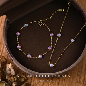 Pode incluir: Um conjunto de colar e pulseira dourados. Cada pe&ccedil;a apresenta pedras preciosas redondas, iridescentes, roxas e azuis. As joias s&atilde;o exibidas em uma caixa de joias marrom. O texto "MICHELLELEE STUDIO Handcrafted Jewellery" &eacute; vis&iacute;vel.