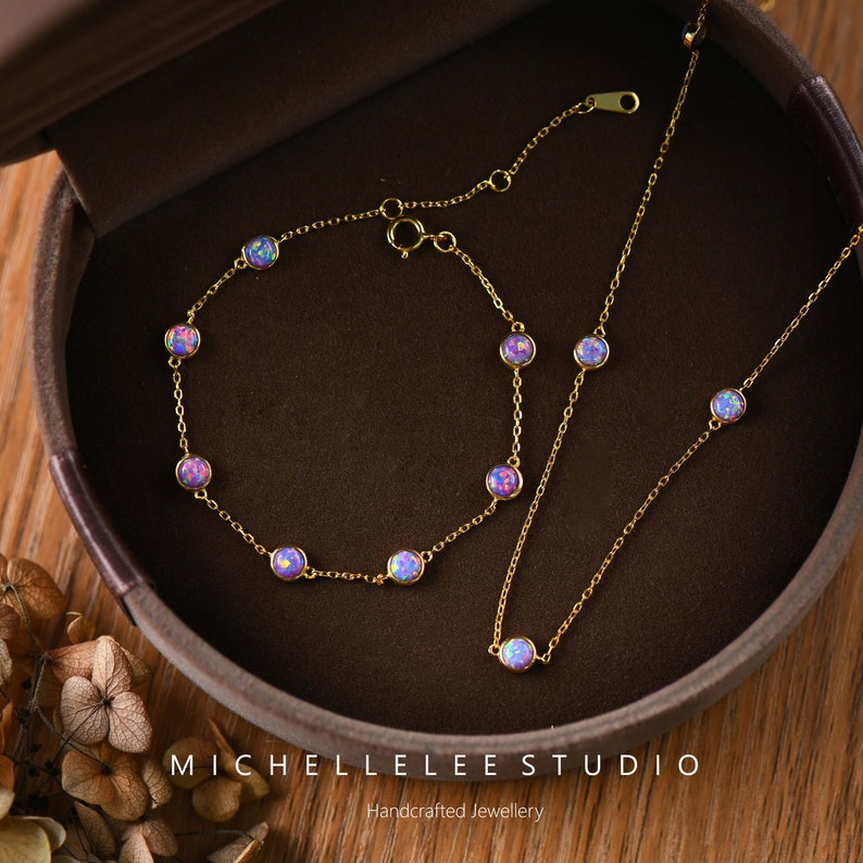 Pode incluir: Um conjunto de colar e pulseira dourados. Cada pe&ccedil;a apresenta pedras preciosas redondas, iridescentes, roxas e azuis. As joias s&atilde;o exibidas em uma caixa de joias marrom. O texto "MICHELLELEE STUDIO Handcrafted Jewellery" &eacute; vis&iacute;vel.