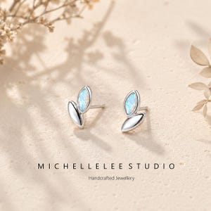 Può includere: Un paio di orecchini a lobo in argento con un design a foglia. Ogni orecchino presenta una forma a foglia in argento lucido con una pietra opalescente ovale. Il testo "MICHELLELEE STUDIO" e "Handcrafted Jewellery" è visibile.