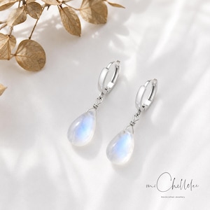 May include: Silver drop earrings featuring pear-shaped moonstone pendants with an iridescent sheen. The earrings have a hinged hoop fastening. The image shows the earrings against a white background. The brand name "mi Chellelee" is visible.