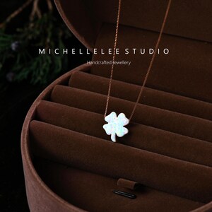 May include: A necklace with a four-leaf clover pendant with an opalescent finish. The pendant is suspended from a fine, rose-gold coloured chain. The jewellery is displayed in a brown velvet jewellery box. The text "MICHELLELEE STUDIO" and "Handcrafted Jewellery" are visible.