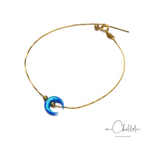 May include: A delicate gold chain bracelet with a blue crescent moon charm. The charm has a shimmering iridescent finish.