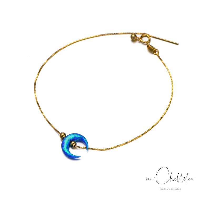 May include: A delicate gold chain bracelet with a blue crescent moon charm. The charm has a shimmering iridescent finish.