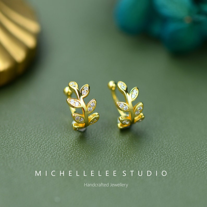 Puede incluir: Un par de pendientes tipo ear cuff dorados con forma de ramas de hojas, cada uno con peque&ntilde;as piedras claras y brillantes incrustadas. El texto MICHELLELEE STUDIO Handcrafted Jewellery aparece impreso en la parte inferior.