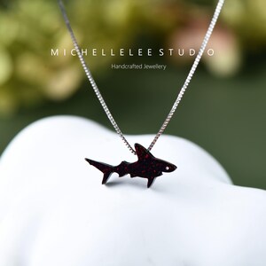 May include: A silver chain necklace with a red glitter shark pendant. The pendant is a silhouette of a shark with a red glitter finish. The necklace is handcrafted jewellery.
