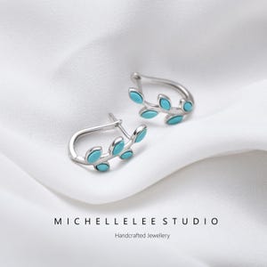 May include: A pair of silver-toned leaf-shaped earrings with turquoise-colored accents. The earrings are displayed on a white, textured surface. The text "MICHELLELEE STUDIO" and "Handcrafted Jewellery" are at the bottom.