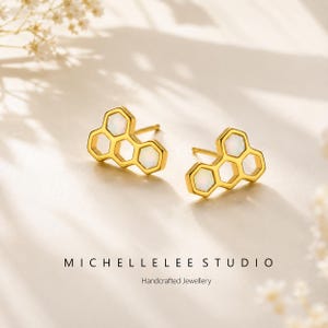 May include: A pair of gold-colored stud earrings in a honeycomb design, each with three hexagonal cells. The cells are filled with white opal stones. The earrings are displayed on a cream-colored surface. The text "MICHELLE LEE STUDIO Handcrafted Jewellery" is below the earrings.