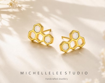 May include: A pair of gold-colored stud earrings in a honeycomb design, each with three hexagonal cells. The cells are filled with white opal stones. The earrings are displayed on a cream-colored surface. The text "MICHELLE LEE STUDIO Handcrafted Jewellery" is below the earrings.