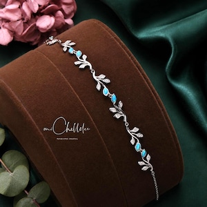 May include: A silver-toned bracelet featuring a vine-like design with alternating clear stones and small, oval-shaped blue opalescent stones. The metal links are shaped like leaves, and the text miChellee Handcrafted Jewellery is visible.