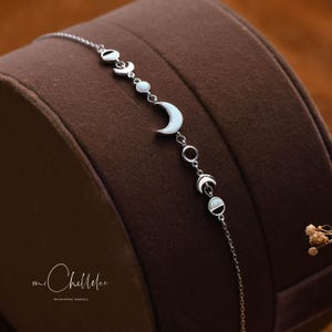 以下が含まれることがあります： 三日月型のチャームとオパールのアクセントが付いたシルバーチェーンブレスレット。ブレスレットは、"mi Chellee handcrafted jewelry"というテキストが書かれた茶色の表面の上に置かれています。