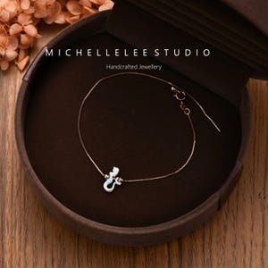 Puede incluir: Una delicada pulsera de plata con un peque&ntilde;o dije blanco en forma de sirena. La pulsera se exhibe en una caja de joyer&iacute;a marr&oacute;n. El texto "MICHELLELEE STUDIO Handcrafted Jewellery" es visible.