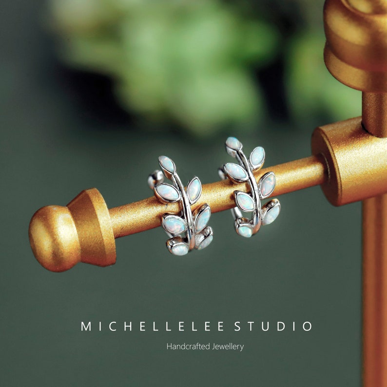 Puede incluir: Un par de pendientes de aro plateados con un dise&ntilde;o de hojas y peque&ntilde;as piedras de &oacute;palo ovaladas e iridiscentes. El texto MICHELLELEE STUDIO Handcrafted Jewellery es visible debajo de los pendientes en el soporte.