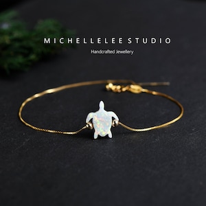 Può includere: Un delicato bracciale a catena dorata con un ciondolo centrale a forma di tartaruga in opale bianco con macchie iridescenti. La tartaruga è affiancata da due piccole perline dorate su una catena sottile. Il testo MICHELLELEE STUDIO Handcrafted Jewellery è visibile.