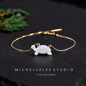 Könnte beinhalten: Ein goldfarbenes Kettenarmband mit einem weißen, opalartigen Schildkrötenanhänger mit irisierenden Sprenkeln. Die Schildkröte wird von zwei kleinen goldfarbenen Perlen auf einer feinen Kette flankiert. Text: MICHELLELEE STUDIO Handcrafted Jewellery.